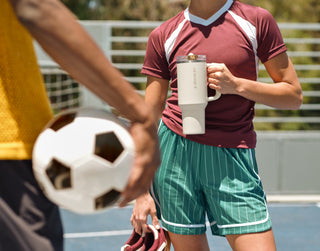 Person in a maroon soccer jersey holding a large white Stanley insulated tumbler on a sunny outdoor field, with another player and soccer ball in the foreground.