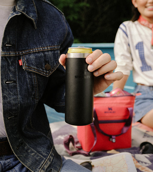 Person in a denim jacket holding a black Stanley can cooler during an outdoor picnic, with a pink cooler bag in the background.