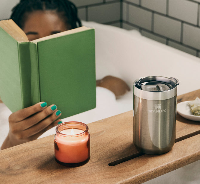 Person reading a green book in a bathtub with a lit candle and a stainless steel Stanley tumbler on a wooden bath tray.