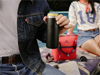 Person in a denim jacket holding a black Stanley can cooler during an outdoor picnic, with a red cooler bag and map in the background.